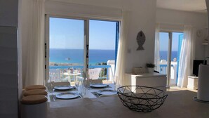 Ein modernes Zimmer mit Balkon und Meerblick, ausgestattet mit einer Essecke und weißen Möbeln sowie einer geometrischen, schwarzen Schüssel auf dem Tisch.