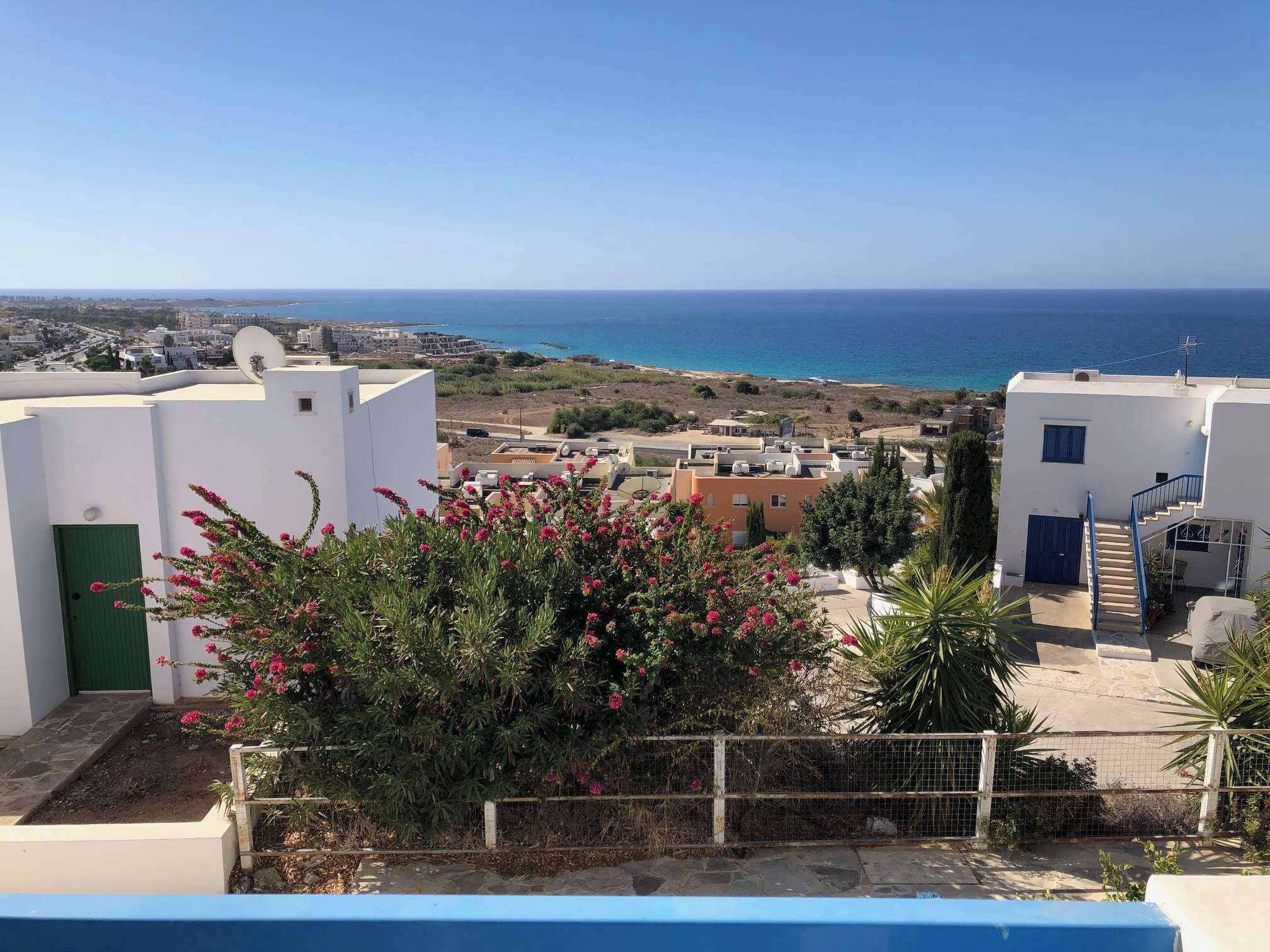 Zona residencial costera con edificios blancos, vista al mar y un jardín con flores rosas.