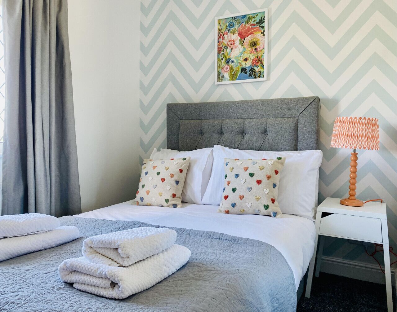 A neatly made bed with white and gray bedding, heart-patterned pillows, and folded towels on a textured gray carpet.