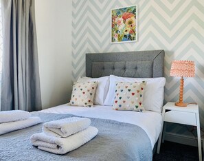 A neatly made bed with white and gray bedding, heart-patterned pillows, and folded towels on a textured gray carpet.