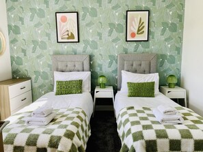 Une chambre d’hôtel avec deux lits, chacun recouvert d’une couvre-lit à carreaux, agrémenté d’oreillers à motifs verts et dotée d’une petite table de chevet avec une lampe.