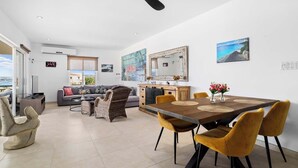 A modern living room with a sofa, dining table, and chairs. There is a kitchen area with a counter and a mirror. The room has a ceiling fan and a view of the outside.