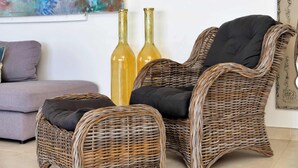 A wicker chair with a black cushion and a matching ottoman, a grey sofa, and two tall glass bottles against a white wall with a colorful abstract painting.