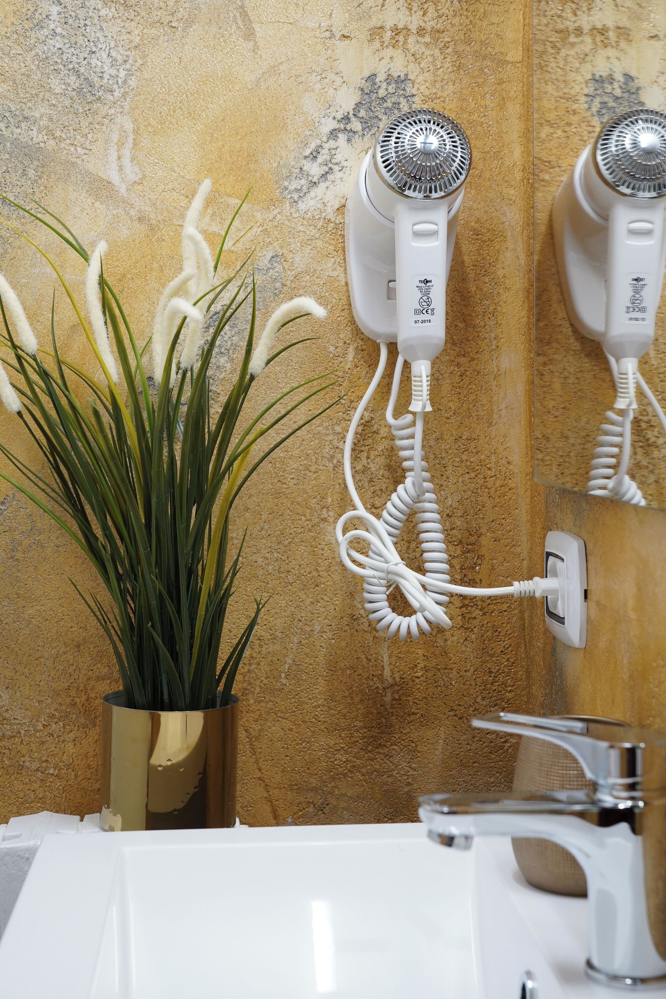 A bathroom with a white sink, a gold vase with green plants, and two wall-mounted hair dryers.