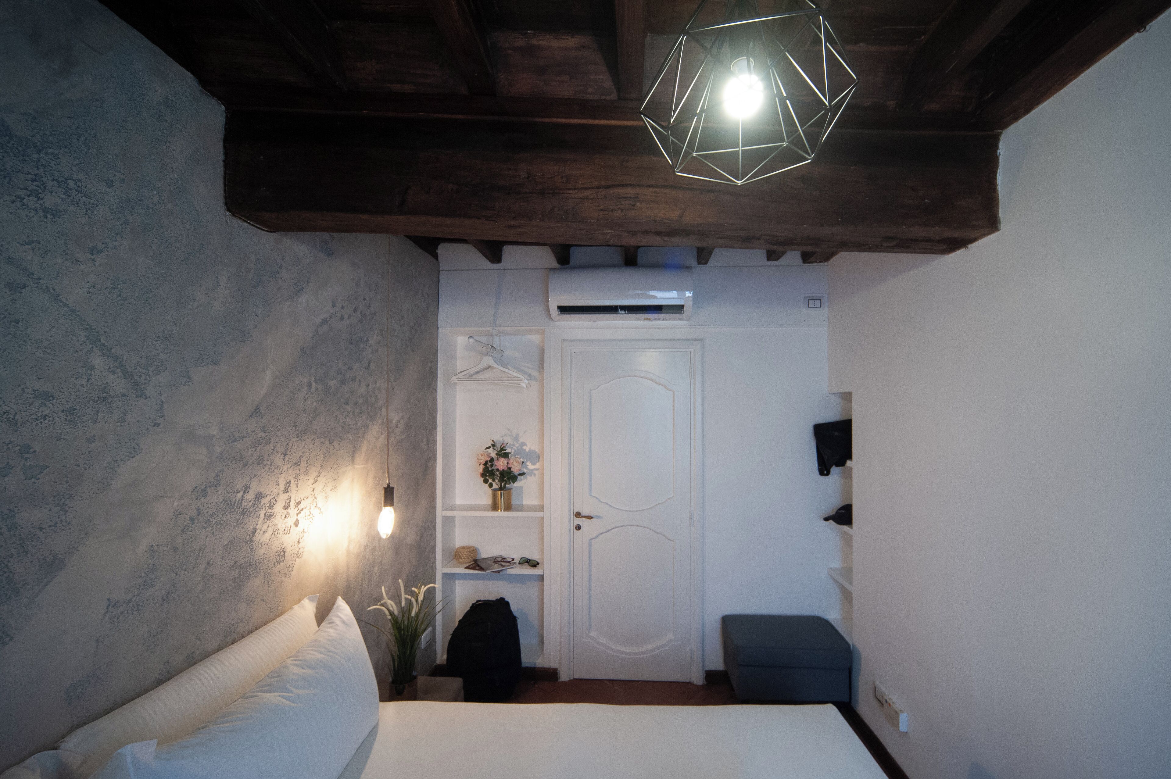 A bedroom with a bed, a hanging light fixture, a white door, and a small shelf with a vase.