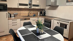 A modern kitchen with white cabinets, a stainless steel range hood, and a built-in oven. There is a dining table with black and white placemats and a vase with flowers.