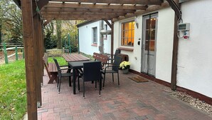 A covered patio with a wooden table and chairs, a white building with a window, and a door.