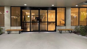 Entrada de un hotel con puerta de cristal, un mostrador de recepción y un banco afuera.