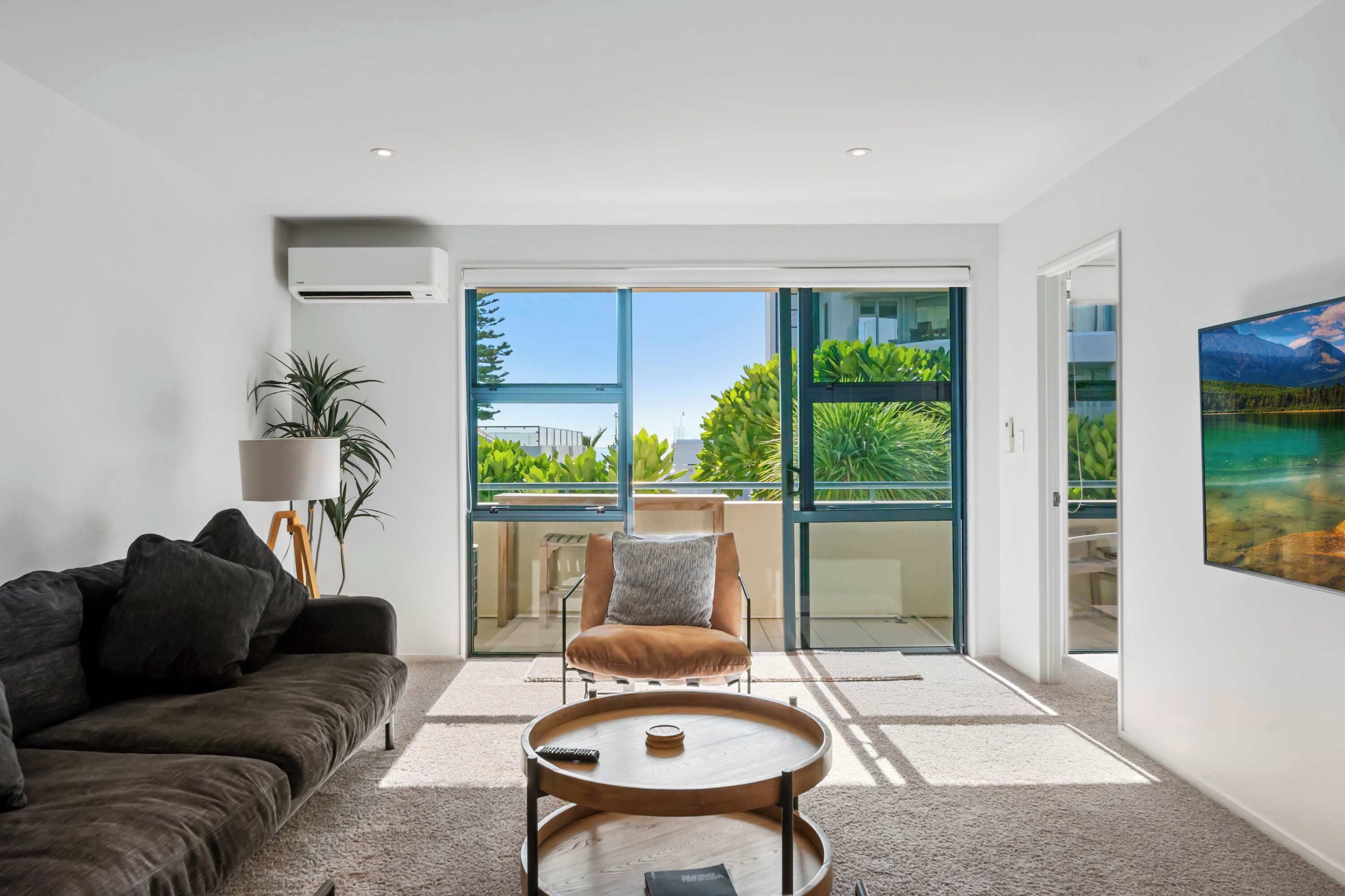 A modern living room with a sofa, a coffee table, a chair, and a large window offering a view of greenery and a building.