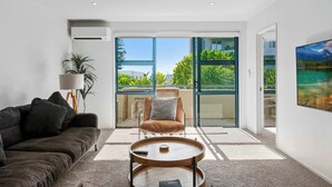 Una sala de estar moderna con un sofá, una mesa de centro, una silla y un ventanal con vistas a zonas verdes y un edificio.