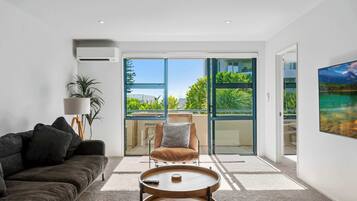 Un salon moderne comprenant un canapé, une table basse, un fauteuil et une grande fenêtre donnant sur un espace verdoyant et un immeuble.