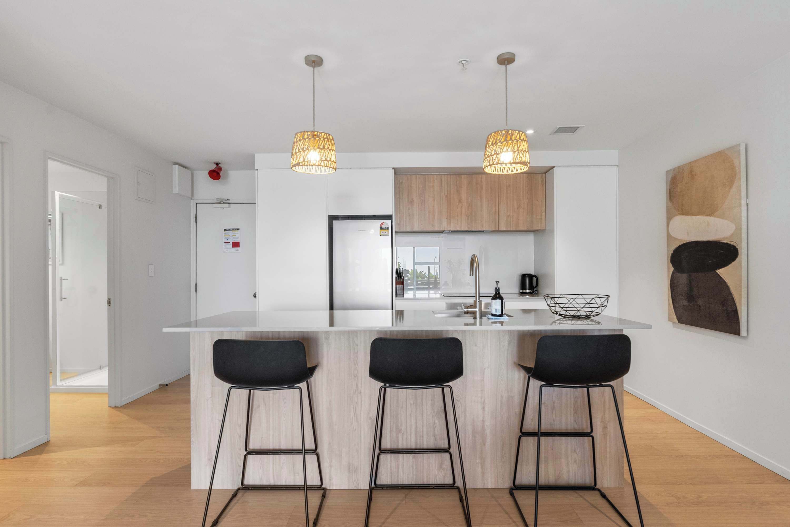 A modern kitchen with a central island, bar stools, pendant lights, and wooden cabinets.