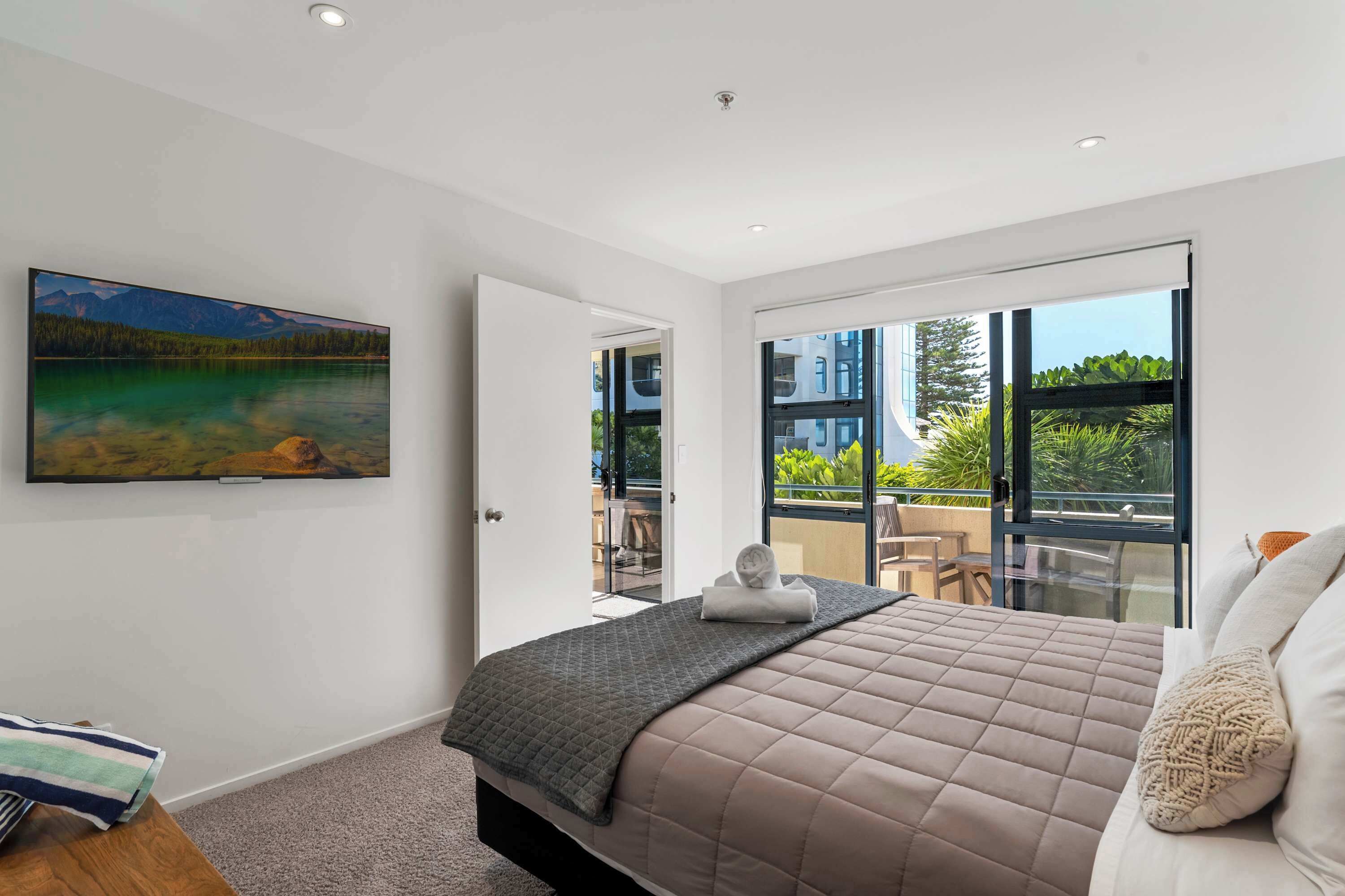 Un dormitorio con una cama grande, un cuadro de un lago y un balcón con vistas a edificios y vegetación.
