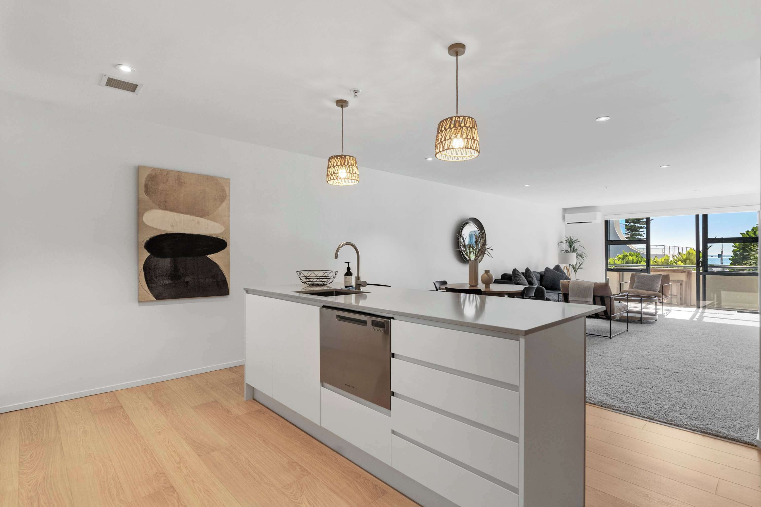 A modern kitchen with white cabinetry, a central island, pendant lighting, and a large window offering a view of greenery outside.