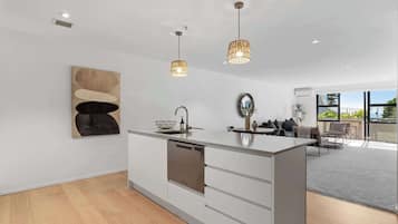 Une cuisine moderne avec des meubles blancs, un îlot central, des luminaires suspendus et une grande fenêtre donnant sur un paysage verdoyant.