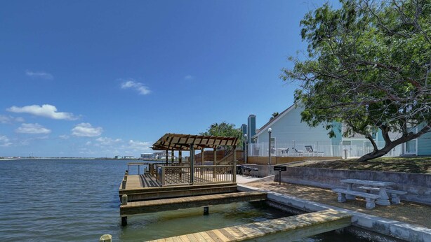 A wooden dock extending into a calm body of water, with a covered seating area and a white building in the background. - Canal Views & Pool | 2 Min to Beach | Fishing Pier (Corpus Christi)
