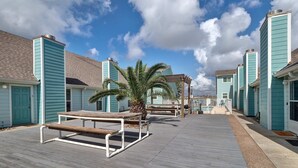 A wooden deck with a bench and a palm tree, surrounded by buildings with teal siding and beige roofs. - Canal Views & Pool | 2 Min to Beach | Fishing Pier (Corpus Christi)