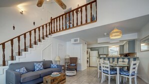 A spacious living room with a grey sofa, wooden coffee table, and a dining area with white chairs and a table. A wooden staircase with a railing leads to an upper level. - Canal Views & Pool | 2 Min to Beach | Fishing Pier (Corpus Christi)