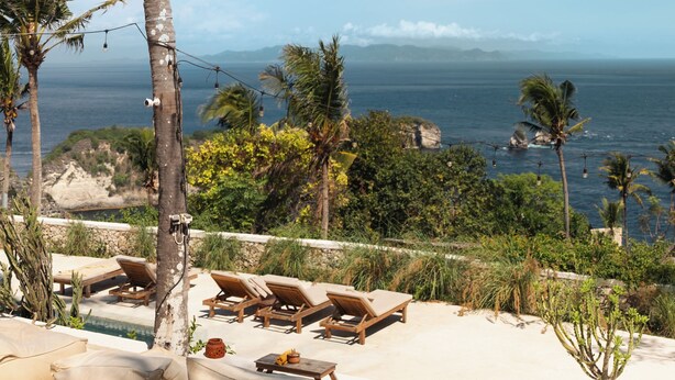 A poolside area with lounge chairs and a small table, overlooking a coastal landscape with palm trees and a view of the ocean. - The Raja Lima (Penida Island)