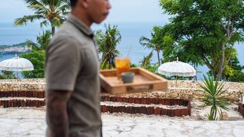A man carrying a tray with a drink and a cup, walking past a stone wall and palm trees with a view of the ocean.