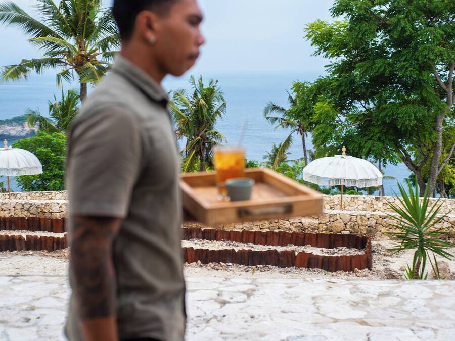 A man carrying a tray with a drink and a cup, walking past a stone wall and palm trees with a view of the ocean.