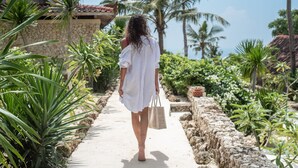 A pathway lined with tropical plants and palm trees, leading to a stone wall and a building with a tiled roof. - The Raja Lima (Penida Island)