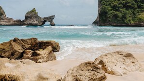 A beach with large rocks, clear blue water, and a rocky cliff with vegetation. - The Raja Lima (Penida Island)