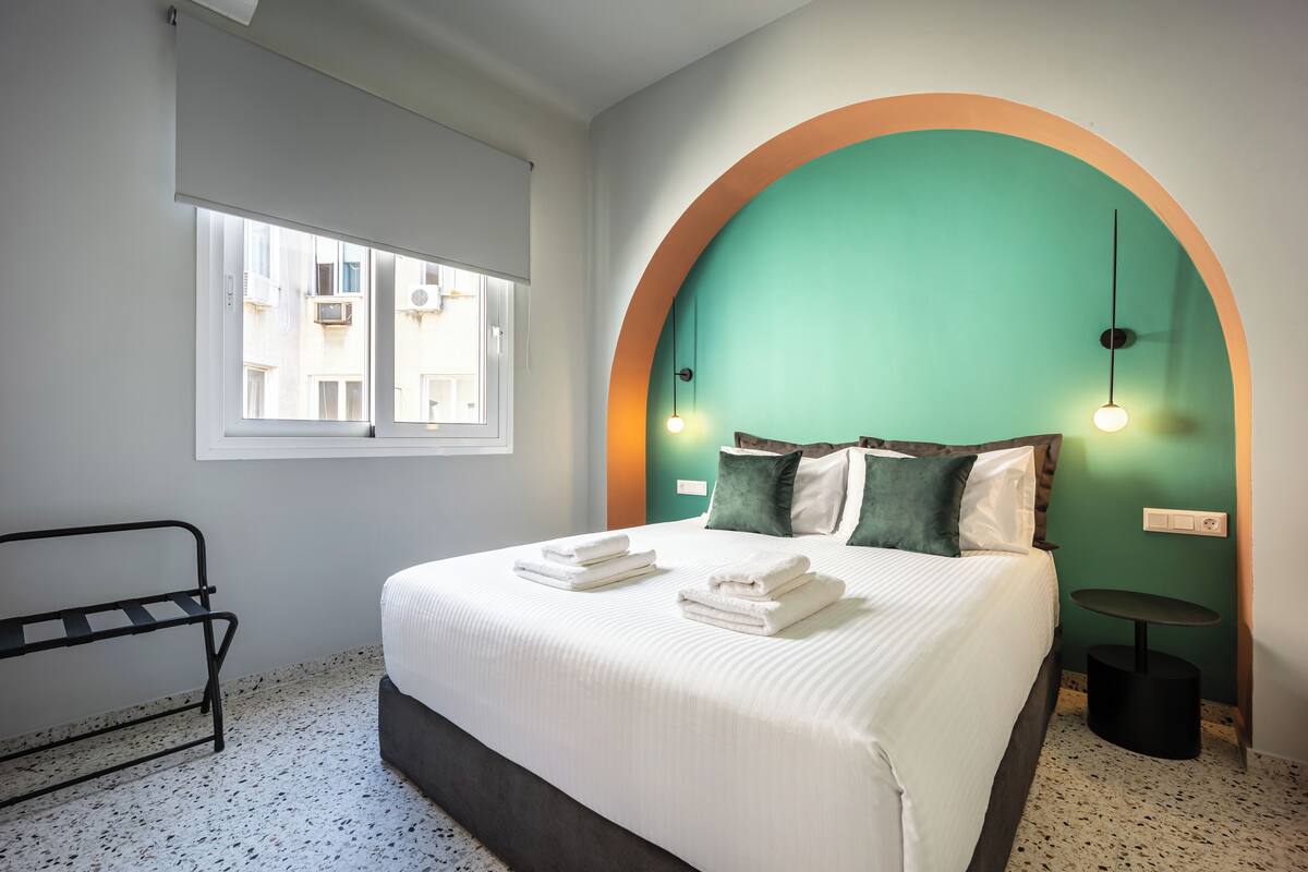 A neatly made bed with folded towels, a bedside table, and a window with blinds.