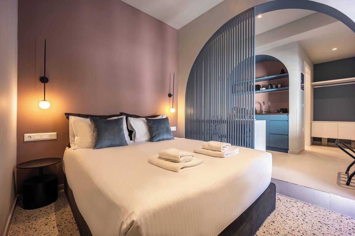 A modern hotel room with a large bed, bedside lamps, a small round table, and a view of the kitchen area.