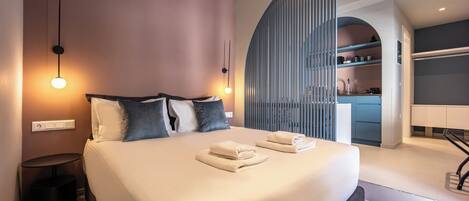 A modern hotel room with a large bed, bedside lamps, a small round table, and a view of the kitchen area.