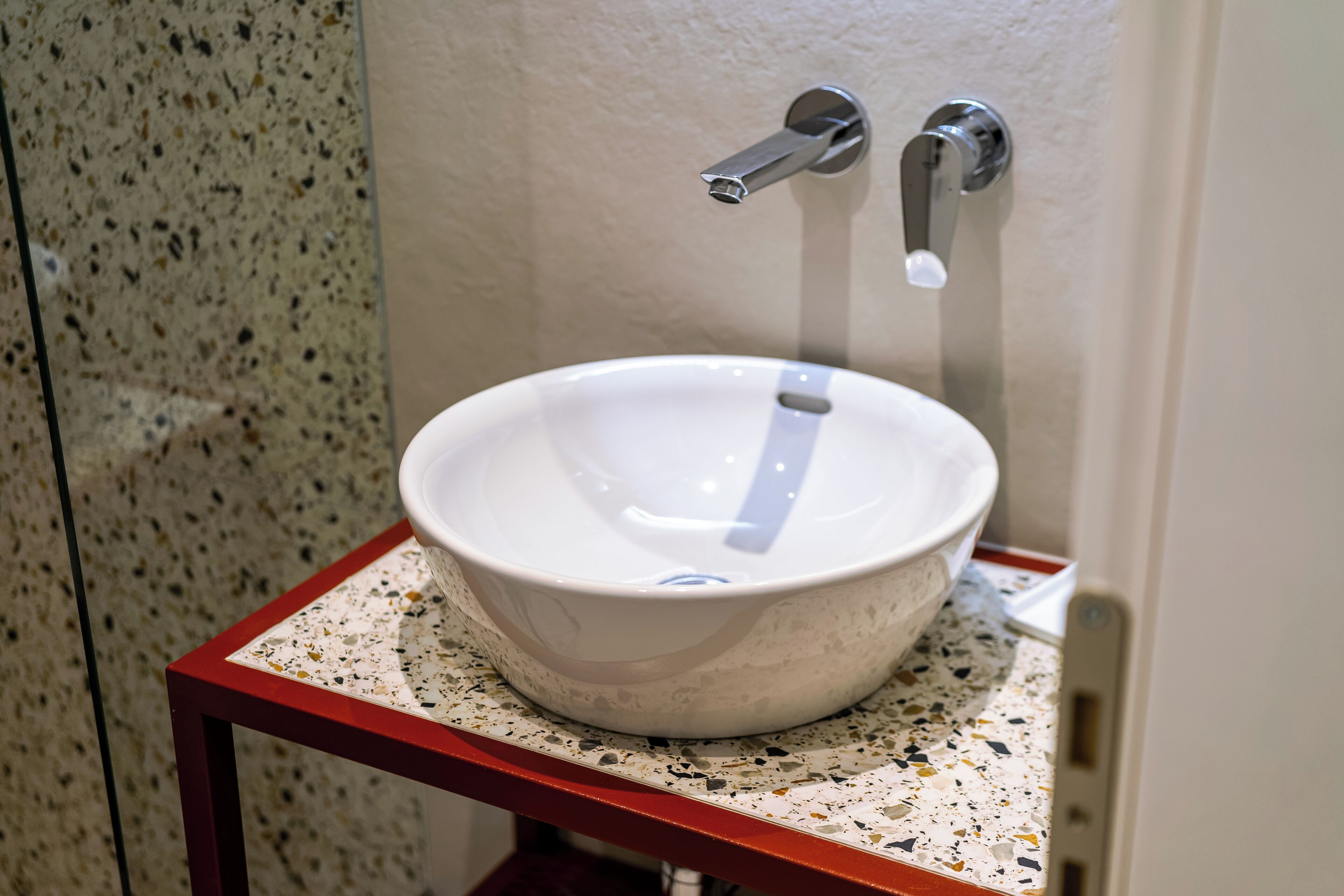 A bathroom with a white ceramic sink on a speckled countertop, a red metal frame, and a wall-mounted faucet.
