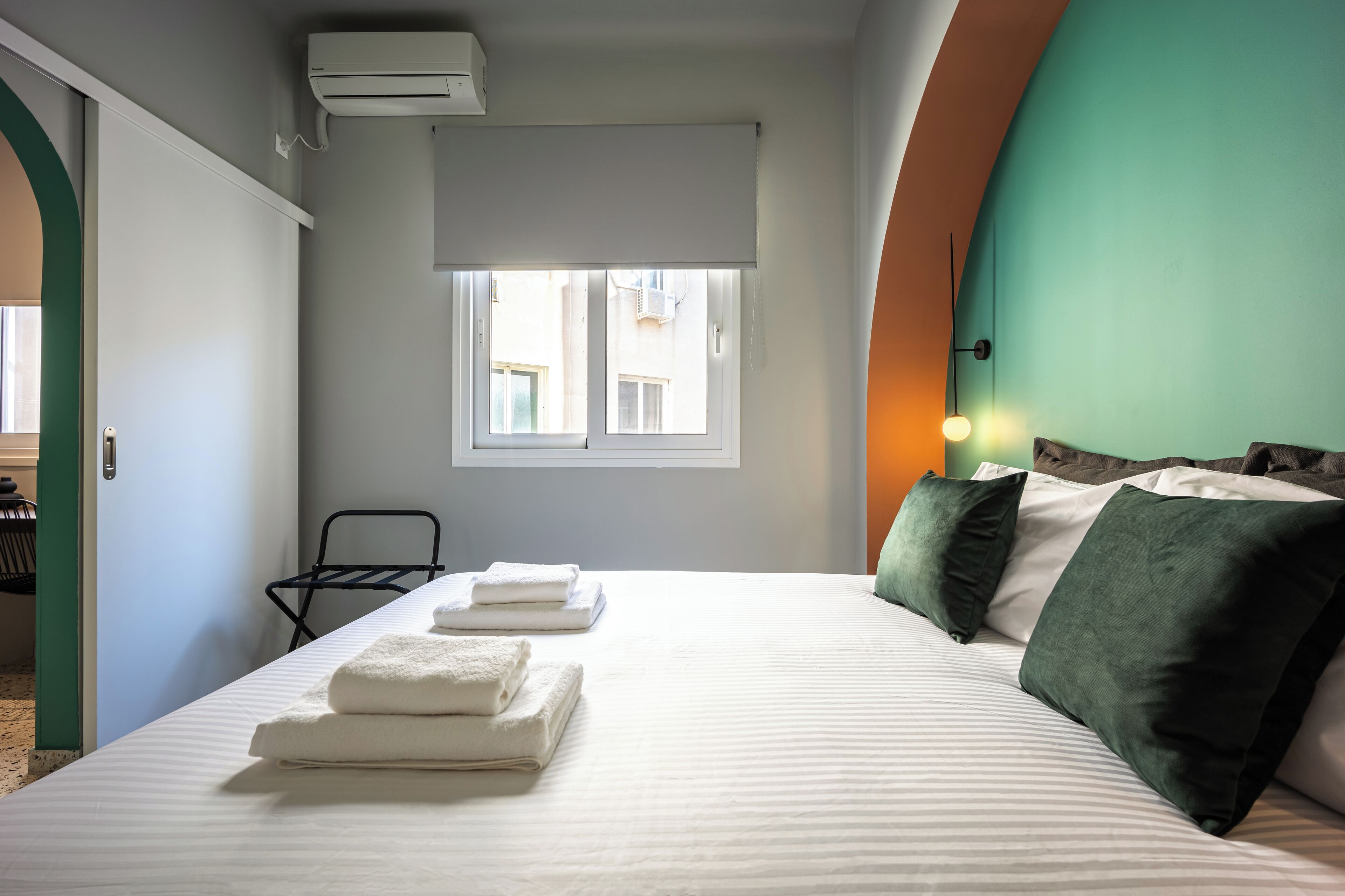 A hotel room with a bed, a window, a chair, and an air conditioning unit.