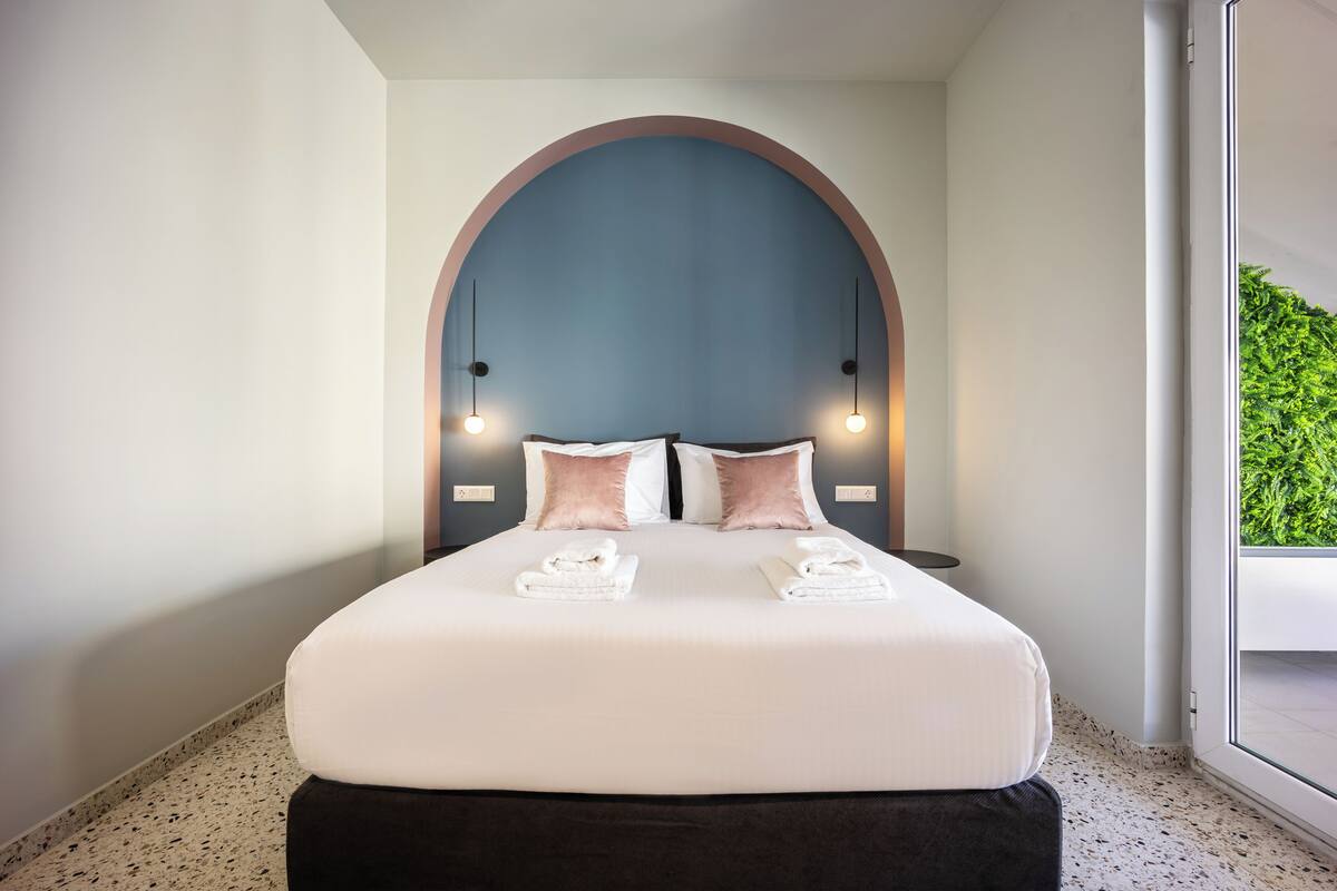 A modern hotel room with a large bed, a unique arched headboard, and a view of greenery through a large window.