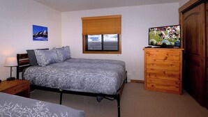 Een slaapkamer met een bed, een dressoir, een televisie en een raam met jaloezieën.