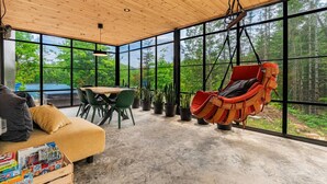 Un interior moderno con techo de madera, ventanas amplias, un sillón colgante, un sofá, una mesa y plantas en macetas.
