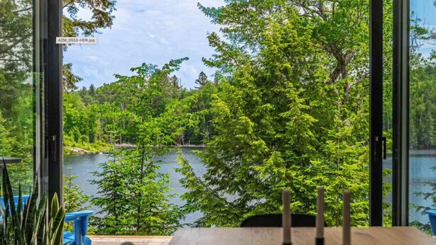 Vista desde una habitación con una mesa y sillas de madera, con vista a un lago rodeado de árboles.