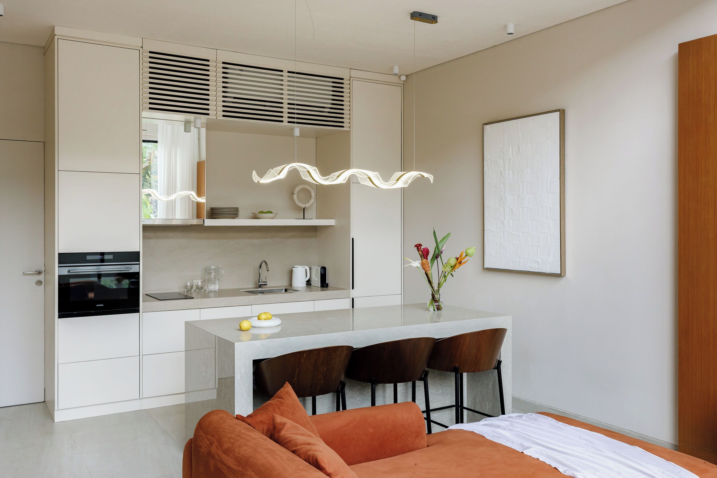 A modern kitchen with a built-in oven, a dining area with a bar stool, and a bed with an orange headboard.