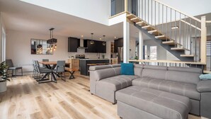 A spacious living area with a large sectional sofa, a dining table with chairs, a kitchen in the background, and a staircase leading to an upper level. - Scandinavian chalet | SPA and Pool Table | Le 70 (Lanaudière)
