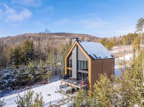 A modern house with a balcony, surrounded by snow-covered trees and a hilly landscape.