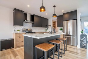 Une cuisine moderne avec des meubles de couleur sombre, un réfrigérateur en acier inoxydable, un îlot central doté de tabourets et un éclairage suspendu.