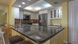 A kitchen with white cabinets, stainless steel appliances, a large island with a sink, and a dining area with chairs. - Beachfront 2B/2B Sandarac Vacation Condo - Million Dollar Gulf View! (Fort Myers Beach)