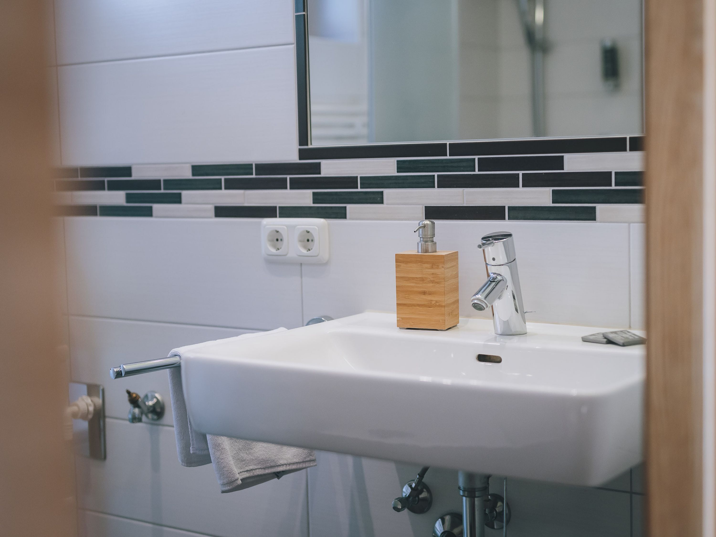 Ein Badezimmer mit einem weißen Waschbecken, einem hölzernen Seifenspender und einem chromfarbenen Wasserhahn. Über dem Waschbecken befinden sich zwei Steckdosen an der Wand.