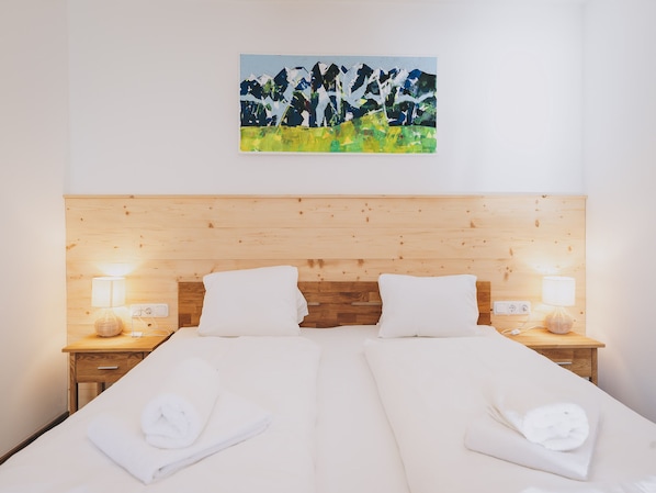 A hotel room with a wooden headboard, two beds with white linens, two bedside lamps, and a painting of a mountain landscape.