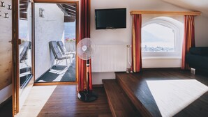 Una habitación con piso de madera, una puerta de vidrio corredera que da a un espacio exterior con una silla y una mesa, un ventilador de pie, un televisor montado en la pared y una ventana con cortinas.