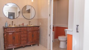 Un baño con tocador de madera, dos espejos redondos, un lavamanos y un inodoro.