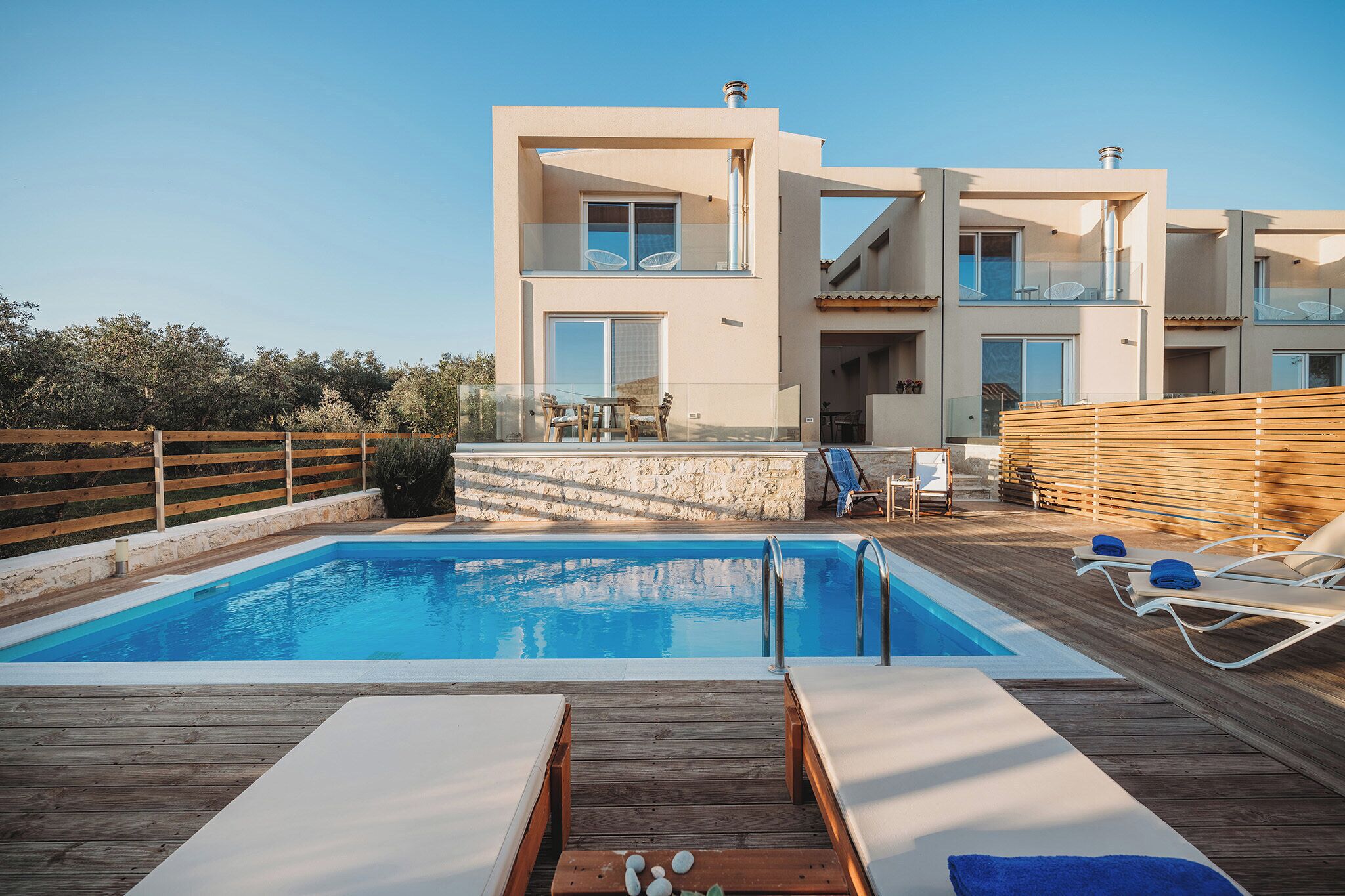 Un edifici modern de dos pisos amb piscina, terrassa de fusta i gandules.