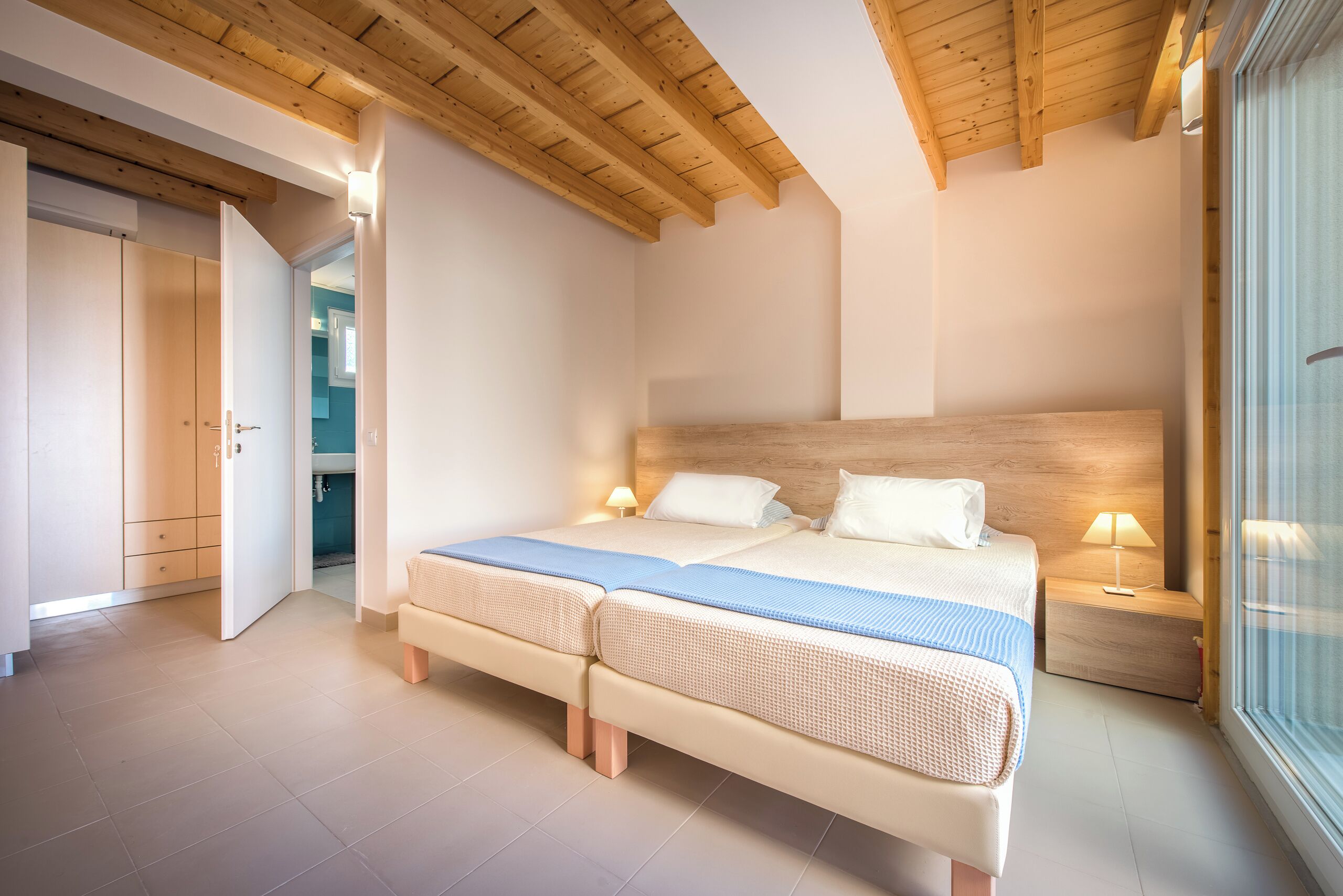 Quarto com duas camas, vigas de madeira no teto e vista para o exterior através de uma grande janela.