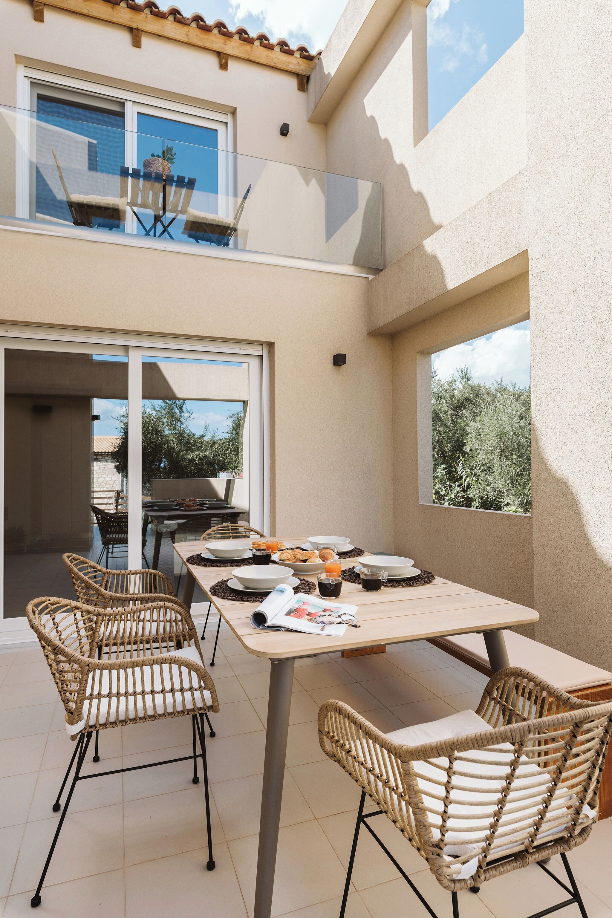 A dining area with a table set for four, surrounded by wicker chairs, and a balcony with a view of trees.