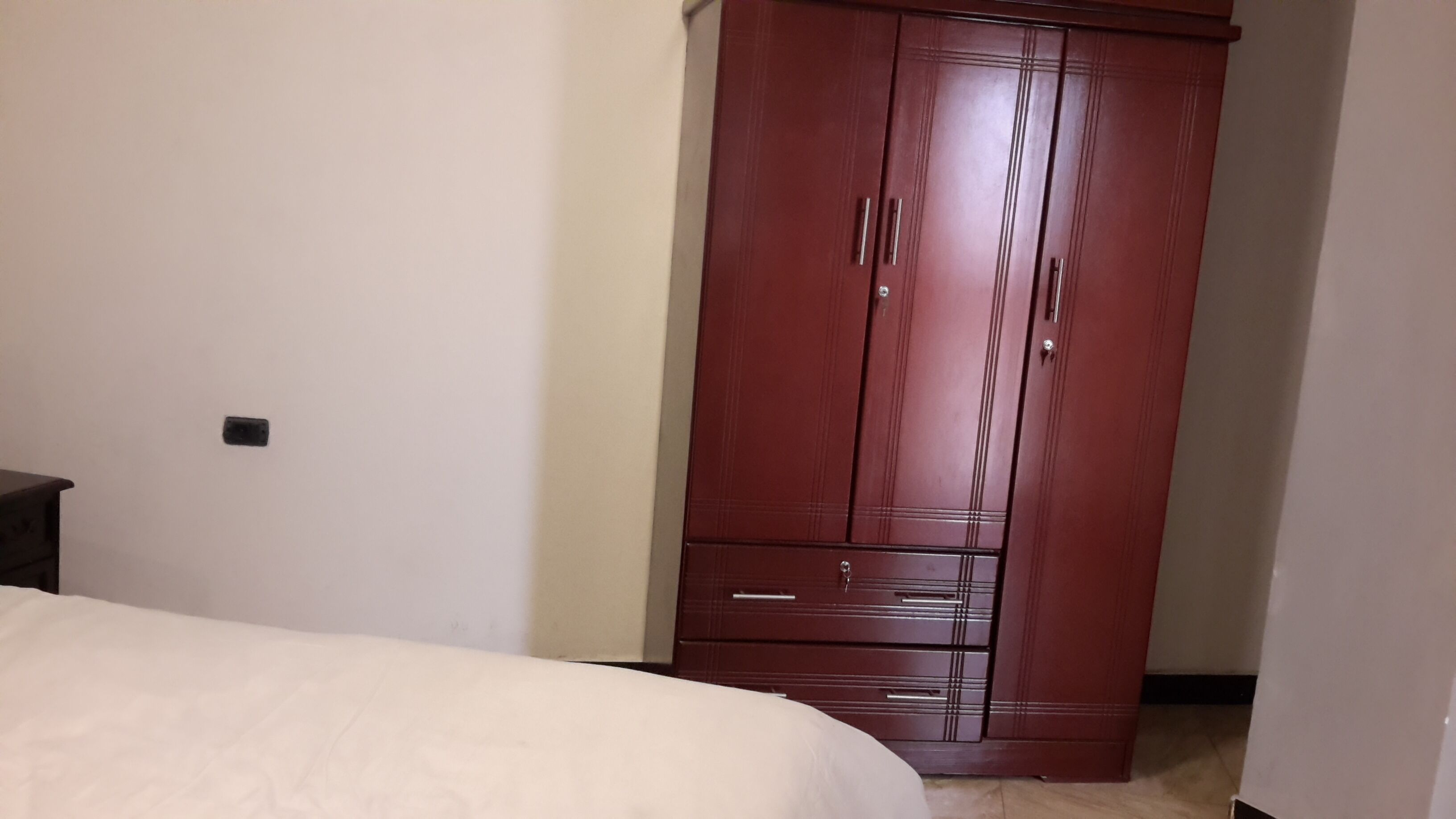 A bedroom with a large wardrobe, a bed, and a small nightstand.