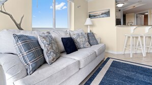 Ein Wohnzimmer mit einer Couch, gemusterten Kissen, einer Lampe, einem Gemälde und Blick auf den Himmel durch ein Fenster.